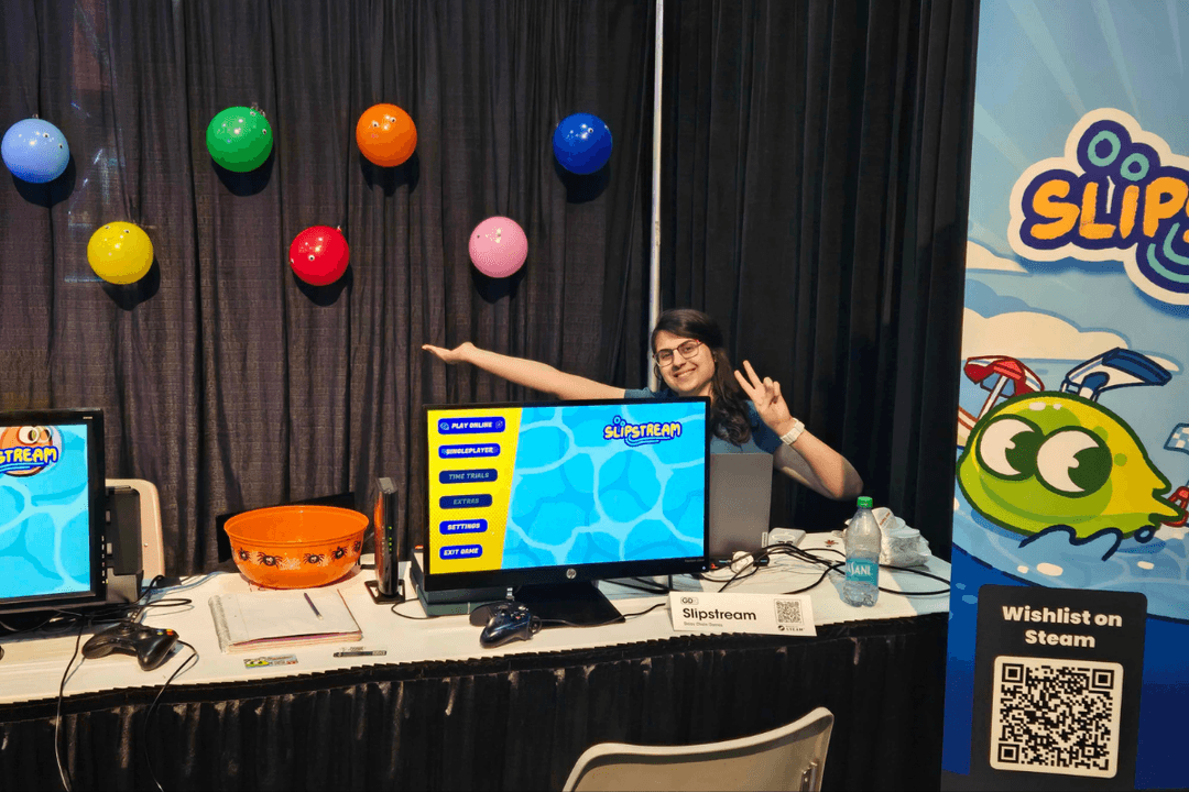 Jawdat Toume, co-founder of Daisy Chain Games, smiles and poses with a peace sign at the Slipstream booth during a gaming expo. She sits behind a table featuring an HP monitor displaying the Slipstream game menu, a game controller, and promotional materials. Colorful character-themed balloons decorate the black curtain backdrop, while a Slipstream banner with the game's green blob mascot and a "Wishlist on Steam" QR code sign are visible to the right.