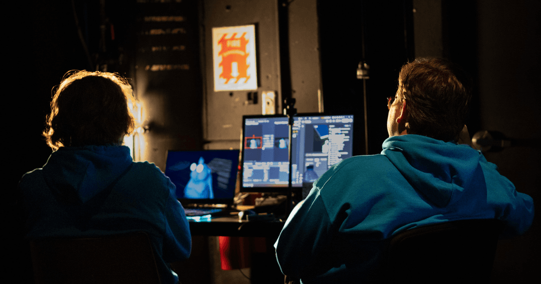 Two people sit at a workstation in a dimly lit studio, working on computers displaying animation or digital media software, photographed during Pixel Blue College’s 2026 AniJam Gala.